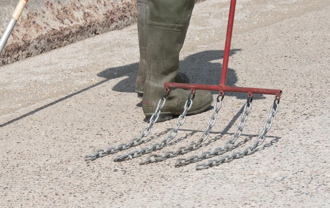 A long red handle with 5 large chains attached across a horizontal bar at the end is dragged across the road surface of a bridge.