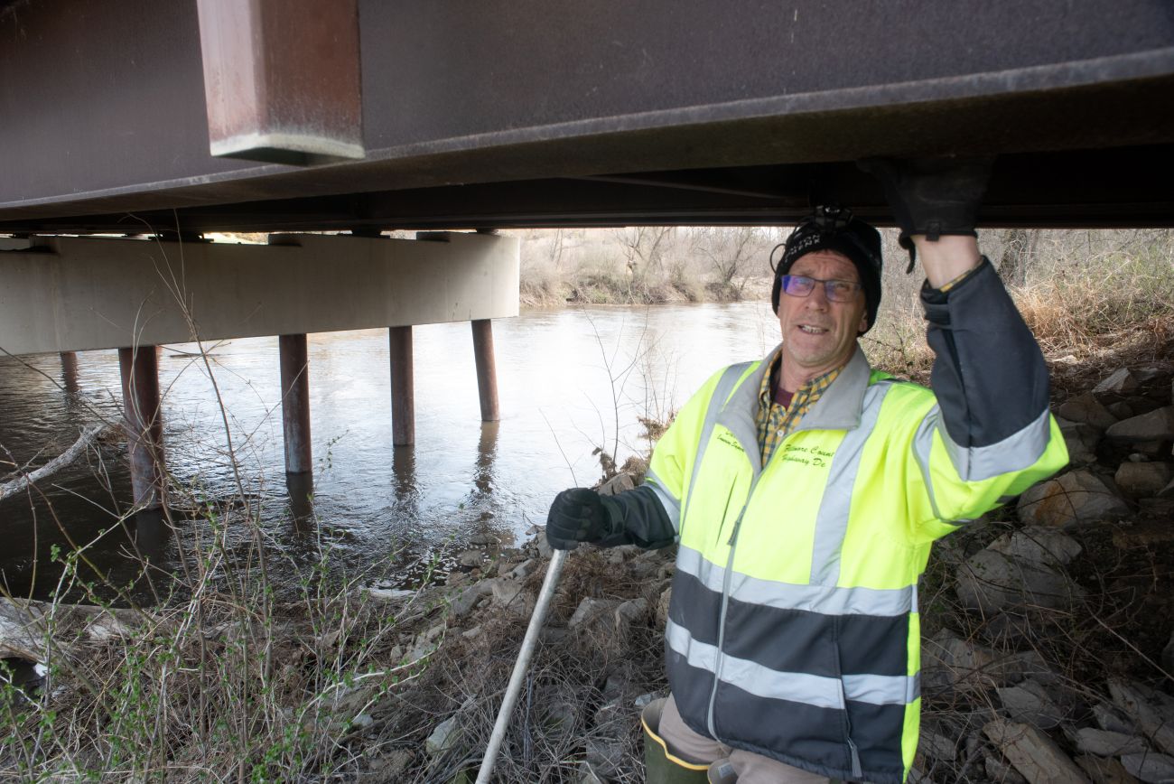 A man in a bright yellow vest with white reflective tape props one hand above his head on the underside of a bridge steel beam.