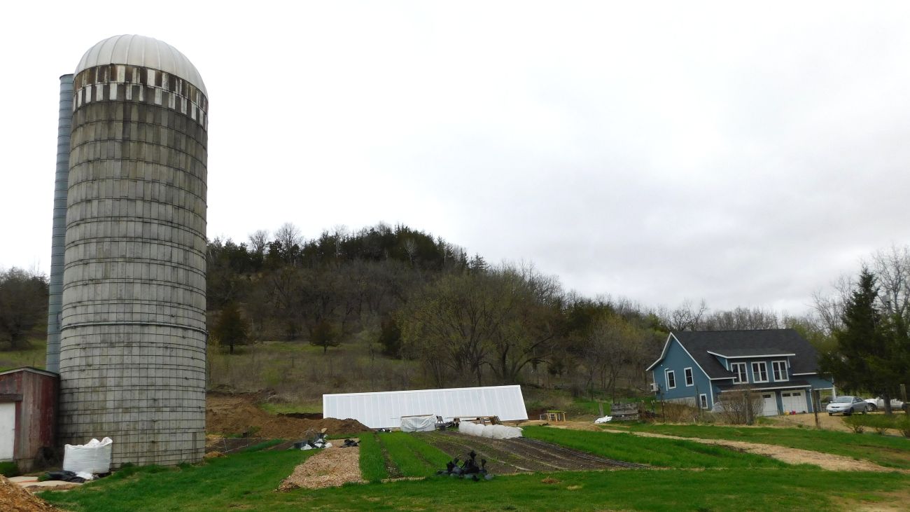 A small farm in front of a wooded hill. On the left is a tall cement silo with a round white top, in the middle is the green house with the white slanted roof, and on the right is a blue two story farm house.