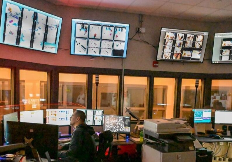 An octagonal room with windows stretching all the way around and large screens hanging above them. At a desk in the middle with another array of screens sits a man in a sheriff's uniform.