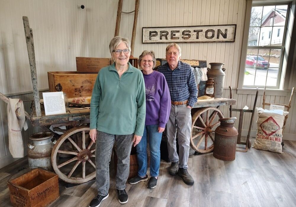 Three older adults stand inside a restored historic railroad-themed museum space in Preston, Minnesota. Behind them is a rustic wooden wagon filled with antique farm and railroad items, including old milk cans, crates, and tools. A weathered wooden “PRESTON” sign hangs on the wall above them, while vintage sleds and historic artifacts decorate the bright cream-colored room with wood plank walls and large windows.