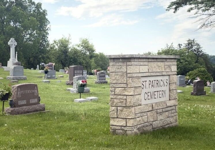 A wide angle shot of a cemetery. In the foreground is a large brick monument with plaque saying St. Patrick's Cemetery. Behind it is green grass and gravestones stretching out and over a small hill. The background is a light blue sky with wispy white clouds.