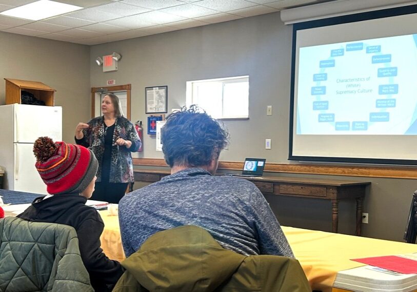 A woman stands at the front of a community meeting room, speaking and gesturing with both hands while facing seated attendees. A projector screen behind her displays a slide titled “Characteristics of White Supremacy Culture” with a circular diagram of terms. Several people sit at long tables in the foreground, listening and taking notes. The room has gray walls, a refrigerator on the left, and daylight coming through a small window.