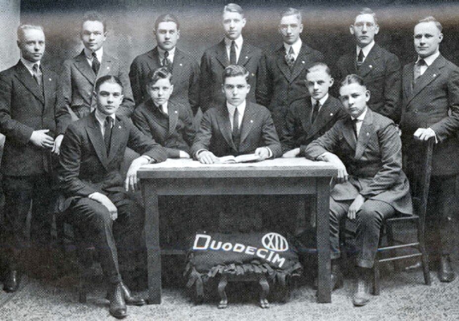 Black-and-white group portrait of twelve young men in dark suits posed around a wooden table, likely a student organization from the early 1900s. A banner reading “Duodecim” rests beneath the table. All are clean-shaven with neatly combed hair, photographed indoors against a plain studio backdrop.