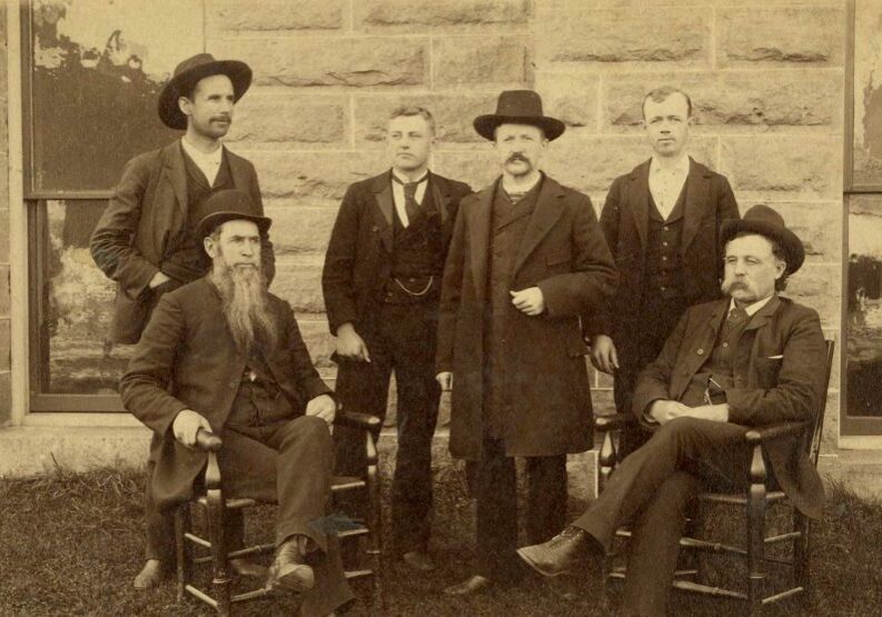 A sepia-toned historical photograph shows six adult men posed in front of a stone building with large rectangular blocks and two windows. Four men stand in the back row while two sit in wooden chairs in the foreground. The men wear late 19th-century clothing, including dark suits, vests, long coats, and wide-brimmed hats. One seated man on the left has a long, full beard reaching down to his chest, while another man in the center has a thick mustache and a dark overcoat, standing with one hand slightly raised. The group faces the camera with serious expressions, typical of formal portraits from that era. The setting suggests a courthouse or civic building.