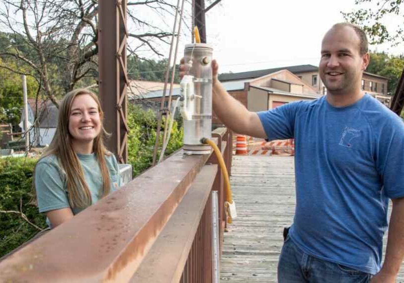 A young woman (left) and a young man (right) stand on either side of a wide, rusted bridge railing and hold a container of water with a rubber tube coming out of the bottom of the container.
