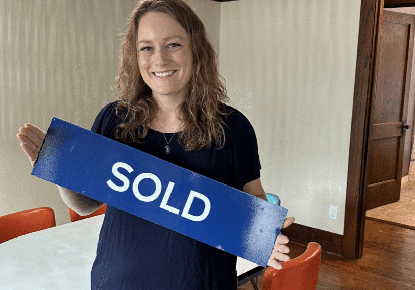 Ayoung woman in a navy blue shirt stands in an older home holding a blue real estate SOLD sign.