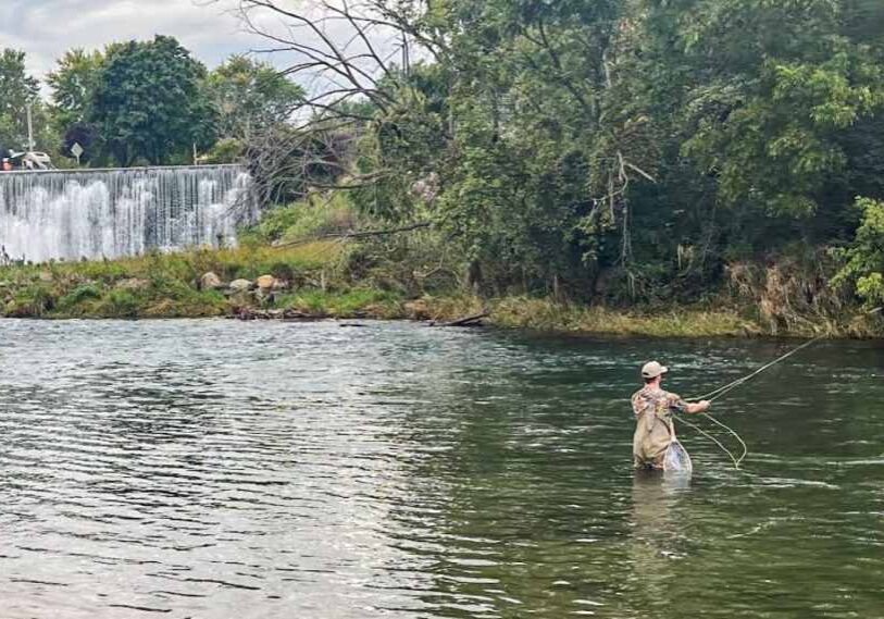 A man in waders stands knee deep in flowing river water just below a short dam. He is casting out a long line from his fishing pole and holding the line in one hand.