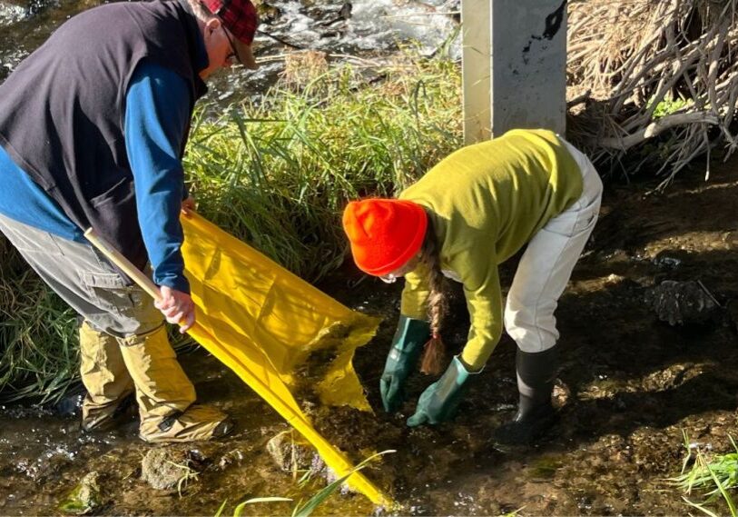 PHOTO 1 Aquatic Guardianship The Importance of Monitoring and M Two people dressed in jackets, hats, and rubber boots stand in shallow water and scoop debris from the bottom of the stream onto a yellow tarp.