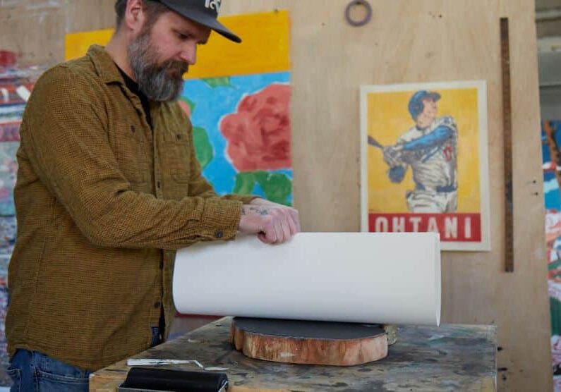 A bearded man wearing a dark baseball cap and a brown button-up shirt stands at a worktable in an art studio. He carefully rolls a large sheet of white paper over a round cross section of a large tree trunk. The workspace is filled with colorful artwork, including a bright poster of a baseball player on the wall behind him. The lighting is warm, and the table shows paint marks and signs of frequent creative use.