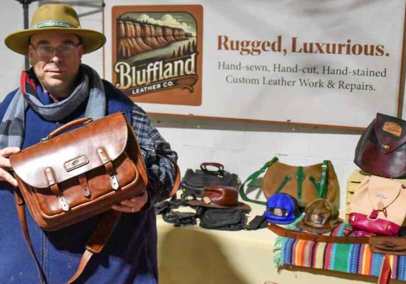 Leather artisan wearing a hat and scarf holds a brown handmade leather messenger bag at a market booth displaying wallets, purses and bags from Bluffland Leather Co.