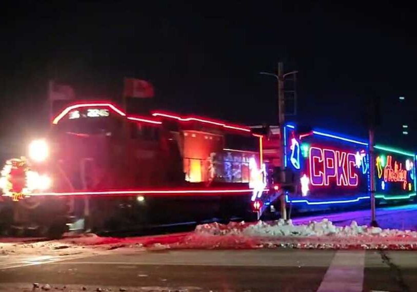 CPKC Holiday Train travels through a snowy crossing at night with the locomotive and rail cars outlined in bright red blue and green Christmas lights.