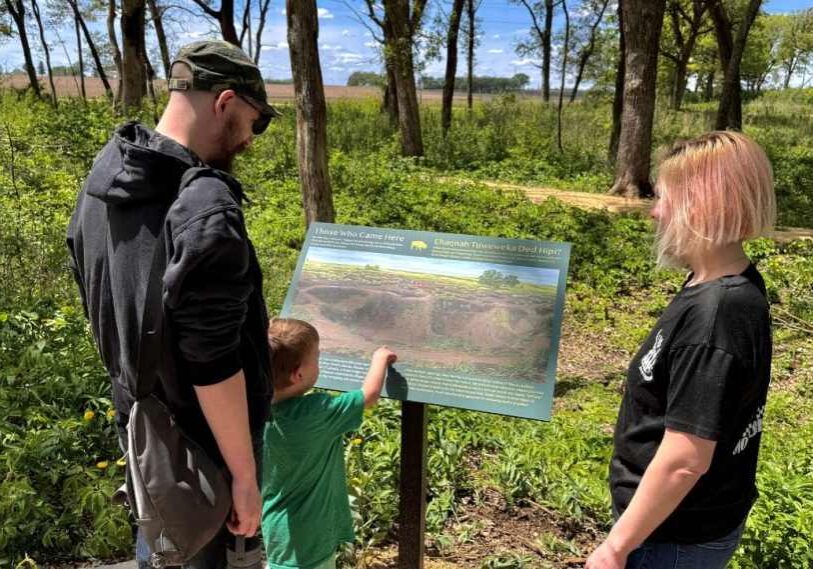 PHOTO 1 Chert Quarry Near Grand Meadow is Rare, Lucky Find for Archaeologists by David Phillips May 2025 A man and a woman and small child stand in a wooded area and look at a large interpretitive map on a stand.