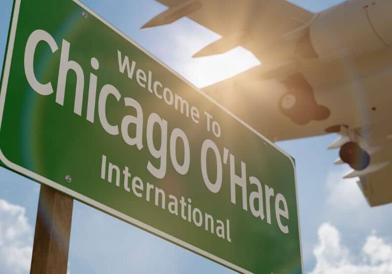 A large green “Welcome to Chicago O’Hare International” sign stands in the foreground while a commercial airplane flies overhead against a bright sky with sun glare and clouds.