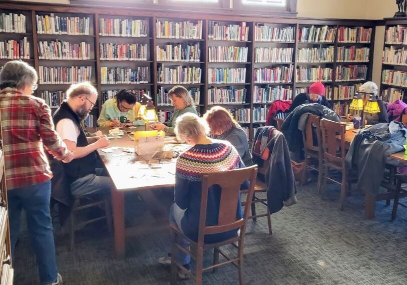 PHOTO 1 Cultivating Community How the Chatfield Public Library is Sowing Seeds by Laurie Byrne March 2025 Groups of people of all ages sit around tables in a library with sun streaming in the windows.. They are leaning over tables sorting garden seeds.