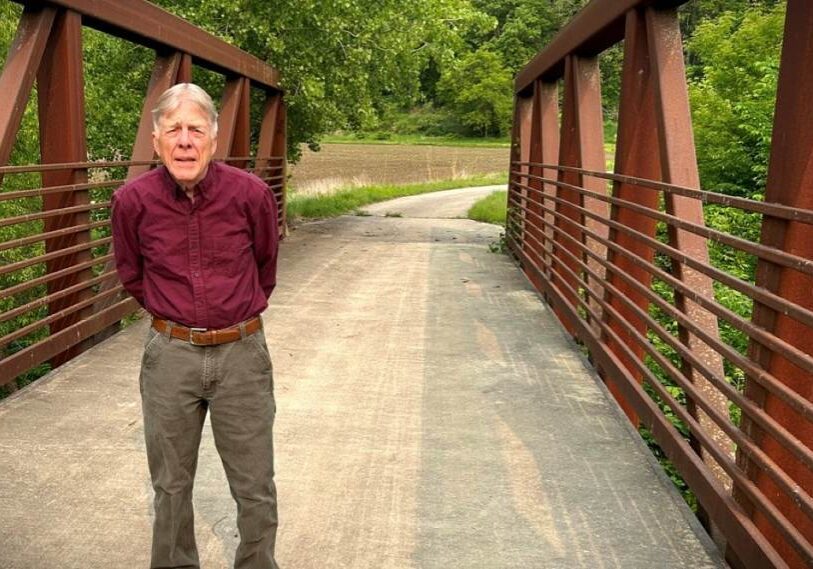 An older man in a maroon long sleeve shirt and tan pants stand in the middle of a small bridge covered in gravel.