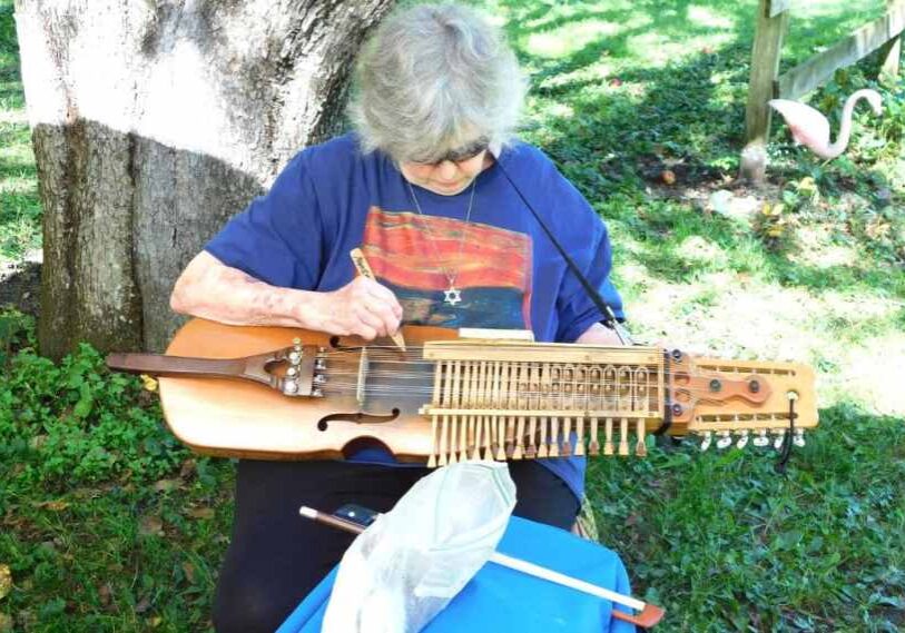 An older woman with gray hair and glasses sits under a large tree on a sunny day, playing a nyckel harpa—a wooden instrument resembling a violin with keys and strings. She wears a blue T-shirt with a colorful abstract print and a Star of David necklace. The scene feels peaceful, with soft sunlight on green grass.