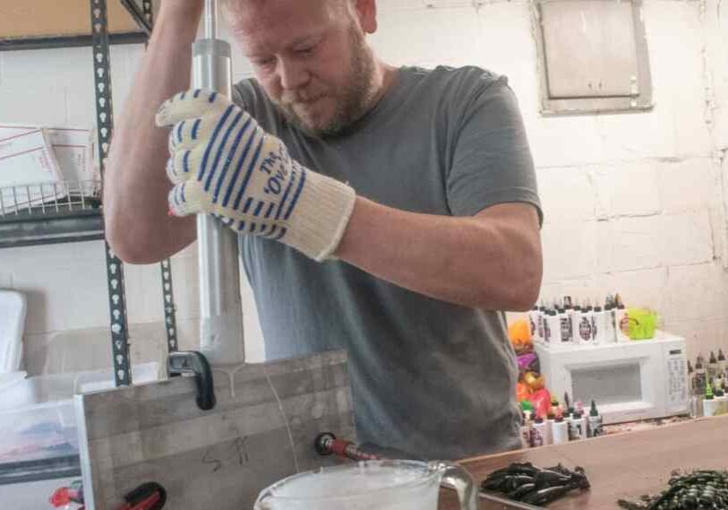 A man wearing a gray tshirt with a large cloth glove on holds a tube sandwiches in a form. His other hand (out of the image) is pushing down on a plunger attached to the tube.