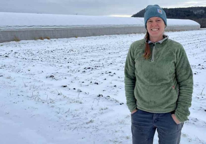 A slender woman in a green zip-up pullover, jeans, and teal stocking cap stands with her hands in her pockets in a snow-covered field beside a long plastic-covered high tunnel greenhouse, with rolling hills in the background on a winter day in southeastern Minnesota.