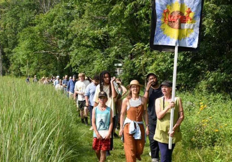 A woman carrying a tall pole with a banner on it is followed by a large crowd of people through a path mown through tall grass.
