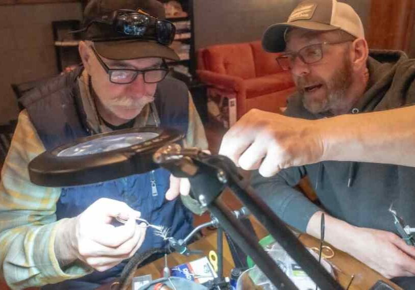 An older man with dark rimmed glasses and dark ball cap intently focuses on tying a trout fly under a bright light and magnifying glass. Another man on the right is speaking and showing him how to do the task.
