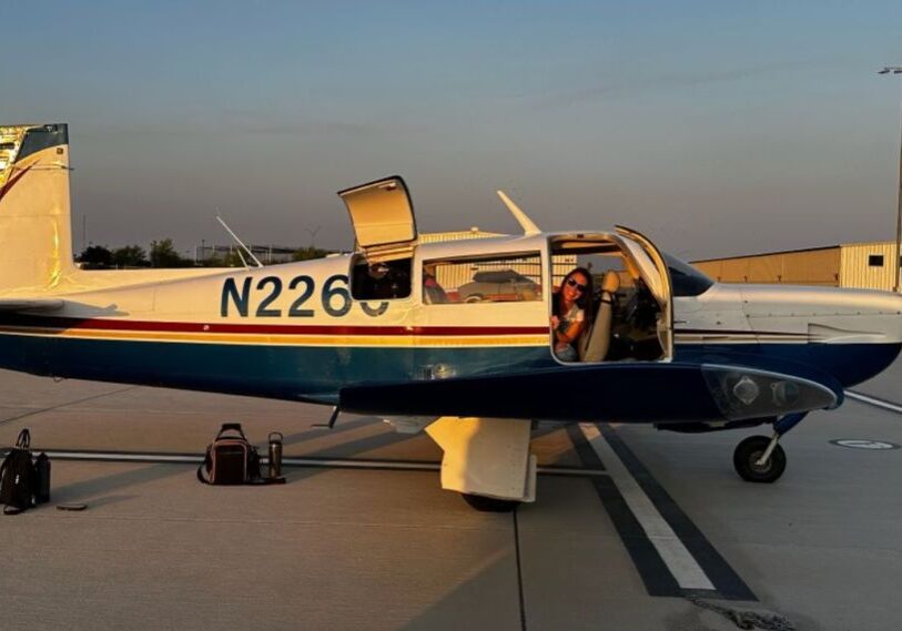 PHOTO-1-Flying-into-Fillmore-County-by-Alison-Leathers-April-2025 A small two-seater white and blue airplane sits on the tarmac with the door open and a women peeking out and smiling.