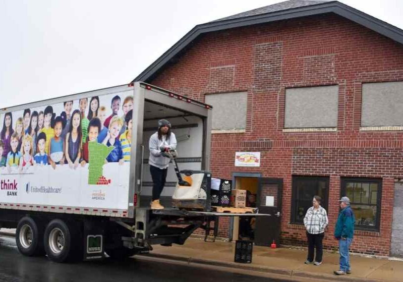 PHOTO 1 Food Shelf by David Phillips October 2025 A large Channel One Regional Food Bank truck is parked beside a red brick building with boarded windows. A worker in a gray hoodie unloads boxes on a lift gate, while two people stand near the open door labeled “Food Shelf.”