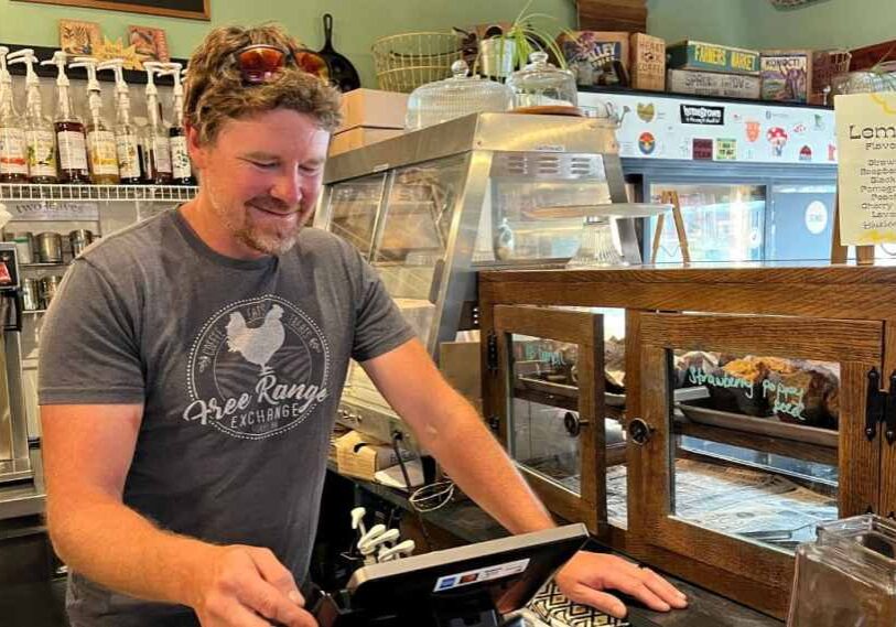 PHOTO 1 Free Range Exchange by Greg Lovell August 2025 A man wearing a dark green tshirt with a logo on the front stand behind a counter and looks down at the register screen.
