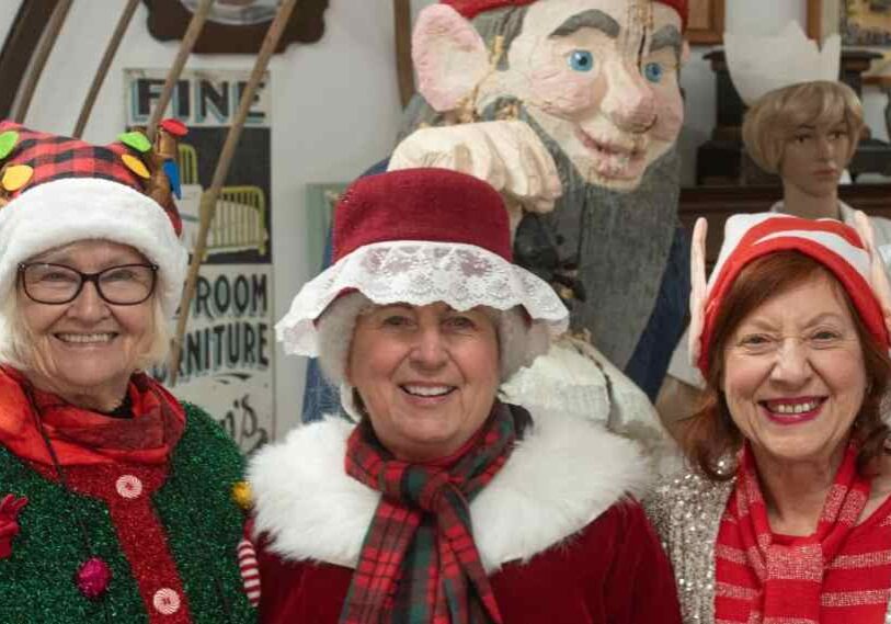 Three older women smile at the camera while wearing festive Christmas hats and scarves inside a museum-like space, with vintage displays and a large carved figure visible behind them.