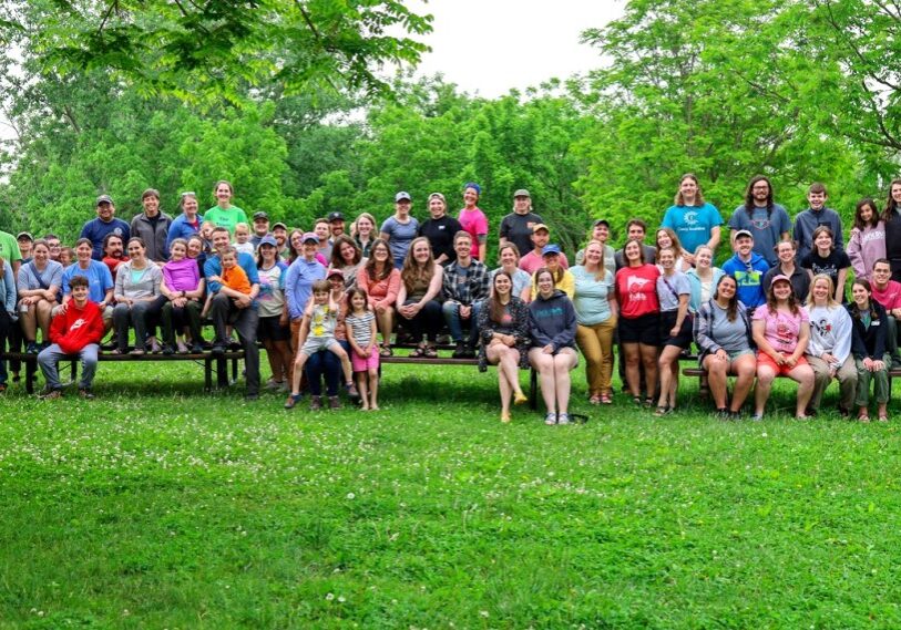 A large group of people of all ages sit on benches in an outside grassy area with leafy trees behind them. All are facing the camera in a group photo setting.