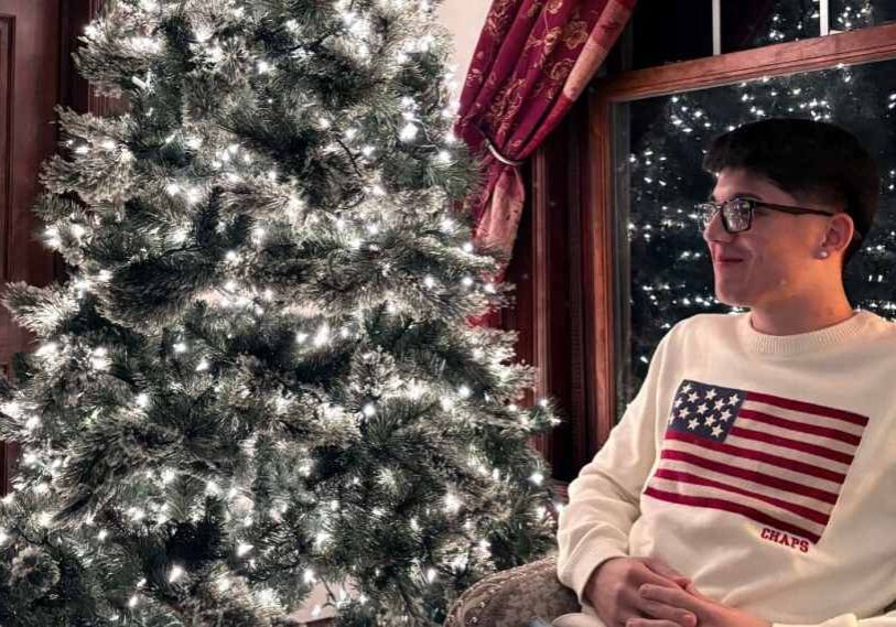 Young man wearing glasses and a white sweater with an American flag sits beside a lit Christmas tree indoors, looking to the side with a calm reflective expression.