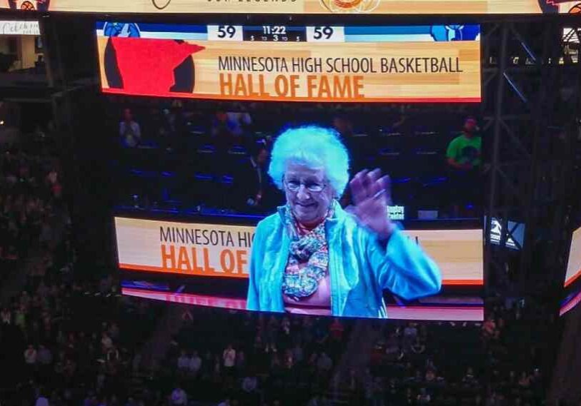 An old women with gray hair is shown on a large jumbotron screen waving at the crowd.