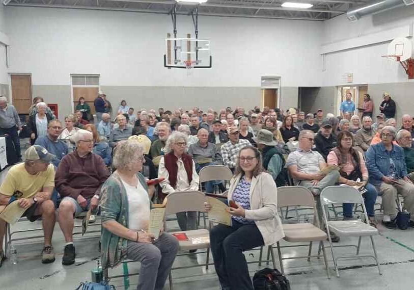 A full gymnasium filled with about 100 people seated in rows of folding chairs for a community meeting. The audience consists mostly of middle-aged and older adults, chatting and holding printed handouts. The room has basketball hoops, beige block walls, and fluorescent lights overhead.