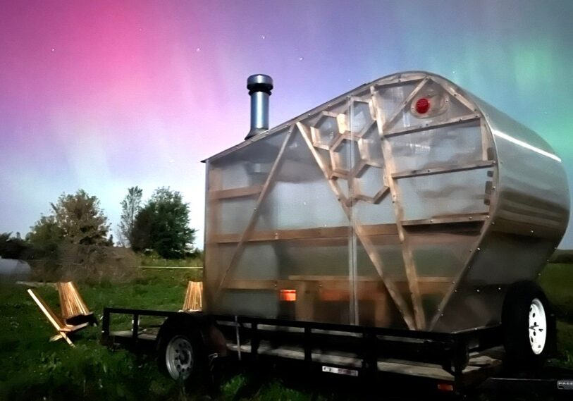 A bright pink, purple, blue, and green sky of Northern Lights shine behind a plexiglas and wood sauna.