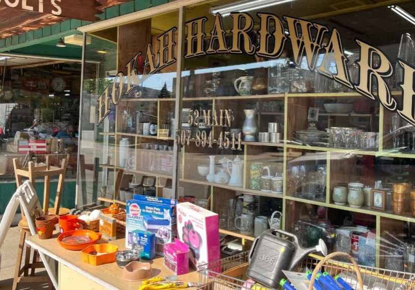 A classic small-town hardware store with a vintage gold-and-black sign reading “Hokah Hardware” painted across large front windows. The display window is filled with household items—glassware, pitchers, and metalware—while tables outside hold brightly colored bowls, tools, and small appliances for sale. The green storefront and wooden overhang evoke an old-fashioned Main Street charm.