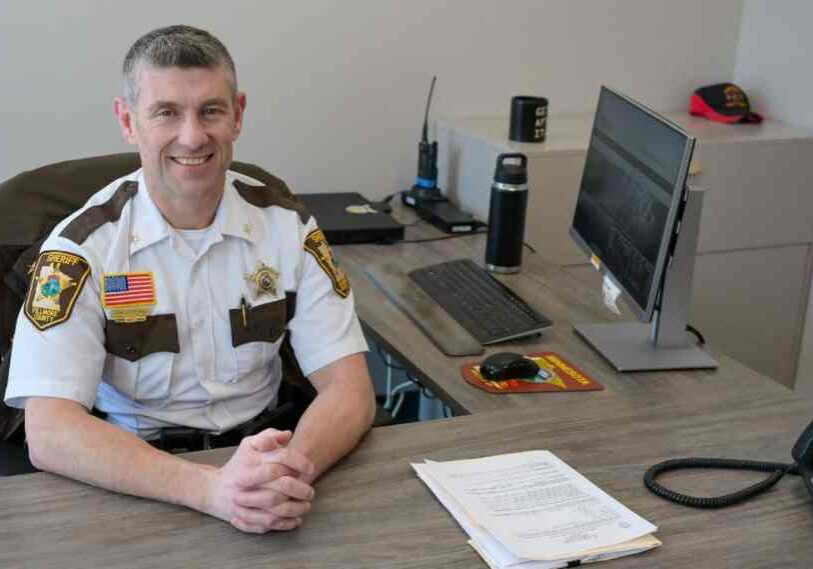 A man with graying hair wearing a white short sleeve shirt with brown pocket flaps, a flag emblem, and a sheriff's star sits at a clean desk and smiles at the camera.