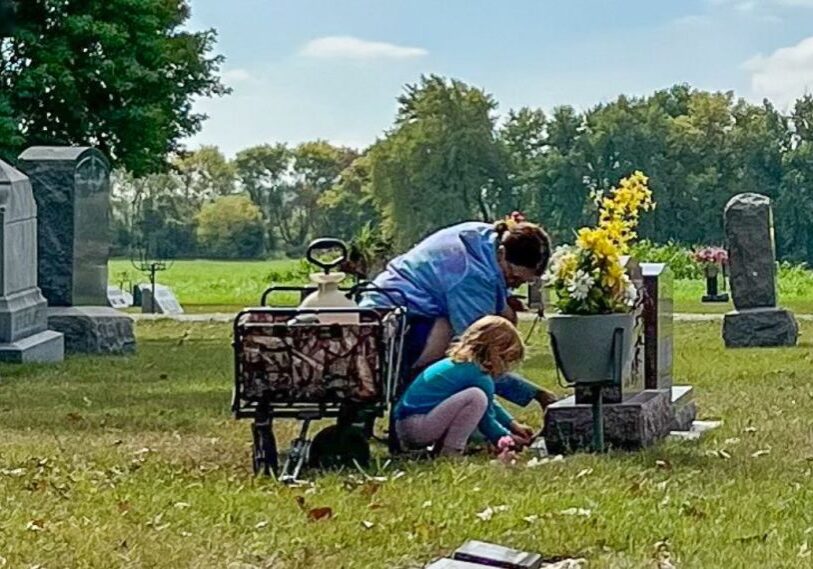 PHOTO 1 Jill Helget and granddaughter clean a headstone A woman and a small child kneel next to a gravestone. Beside them is a wagon holding cleaning supplies. Grass is green, sky is blue.