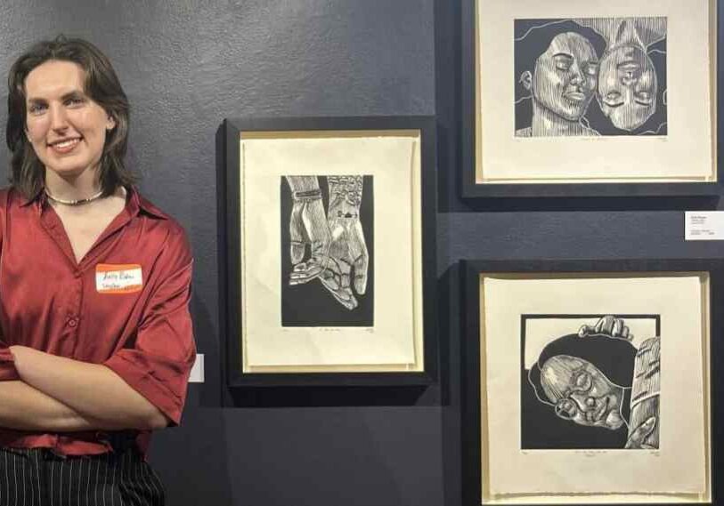 PHOTO 1 Kelly Ristau Hero Image Artist standing beside a series of framed black-and-white linocut prints on a dark gray gallery wall. The artist wears a deep red silk shirt and striped pants, smiling with arms crossed. The prints feature expressive portraits and detailed hands, showing strong linework and emotional realism. Exhibition lighting highlights both the artwork and the artist’s face.