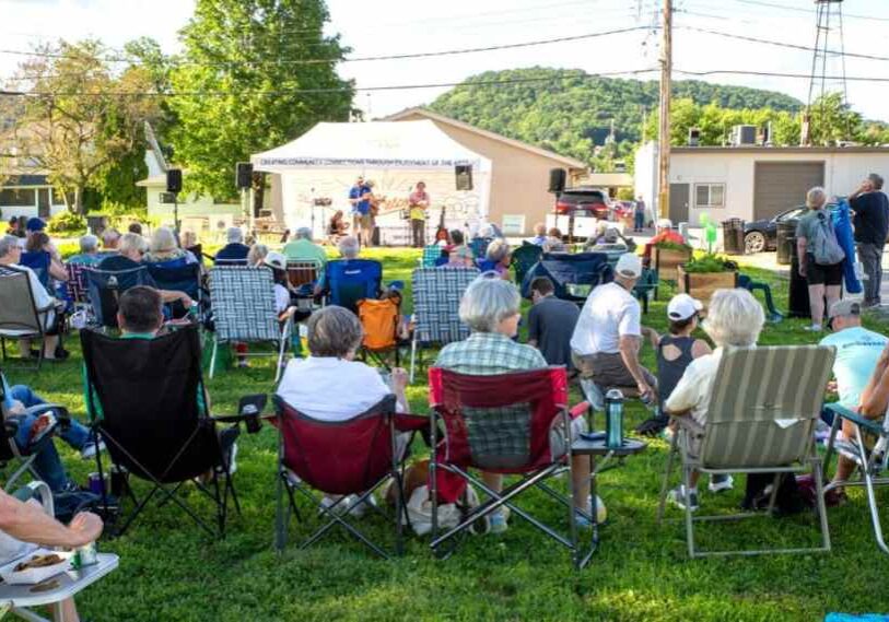 A group of people sit outside in folding chairs on green grass all facing forward watching a musical performance.