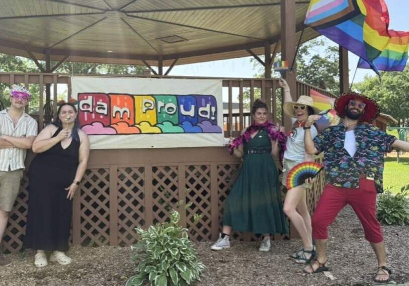 Five people holding photo props stand on either side of a rainbow striped banner with the words "dam proud". The banner is hung on the side of a tall pavilion in a park.