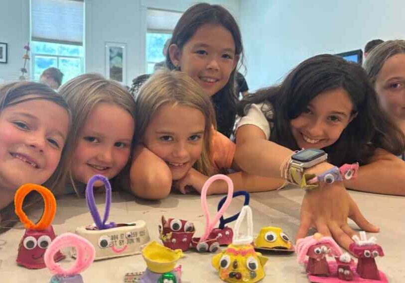 Young children lean on a table with crafts made from egg cartons and pipe cleaners.