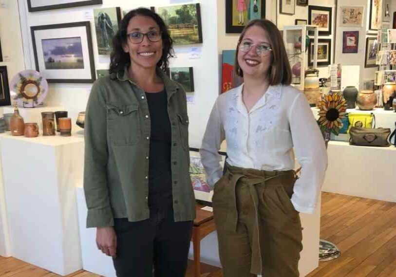 Two women stand in an art studio with pottery and paintings in the background.