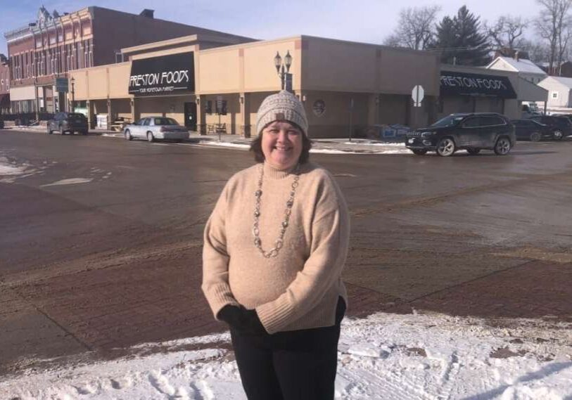 Person wearing a knit hat and beige sweater stands on a snowy sidewalk in a small Minnesota downtown, with a local grocery store and historic brick buildings in the background.