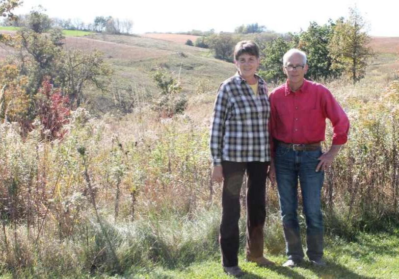 Two people, Barb and Joel Mielke, stand in front of rolling prairie and oak savanna on their Fillmore County property, smiling in the sunlight.