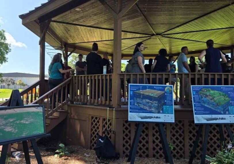 A group of people gather on a large, elevated, circle structure covered by a roof. Signs with writing and images are positioned around the entrance stairway to the gazebo.