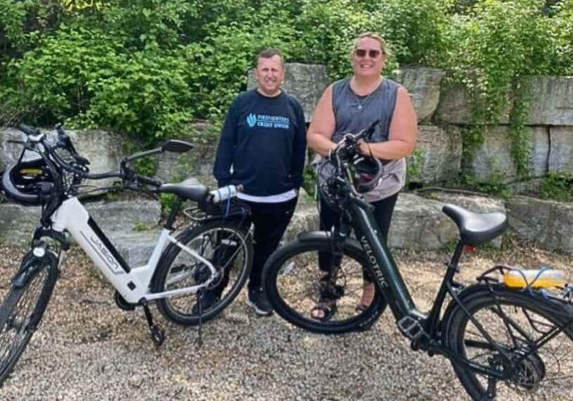 PHOTO 1 “On Your Left!” E-Bikes and the Root River Trail by Steve Harris May 2025 Two people dressed in biking gear stand behind their e-bikes on an outdoor trail.