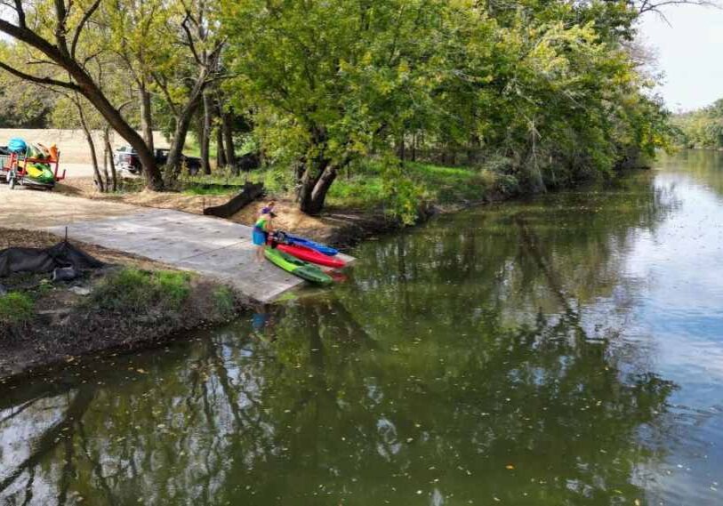 A person wearing a bright green shirt and blue shorts stands beside three colorful kayaks—red, blue, and green—resting on a new concrete boat ramp that slopes gently into a calm, tree-lined river. Two white pickup trucks are parked nearby, one with a small trailer stacked with more kayaks and orange life vests. Sunlight filters through leafy trees, casting dappled shadows on the water, while the opposite riverbank reflects green and gold tones of early autumn.