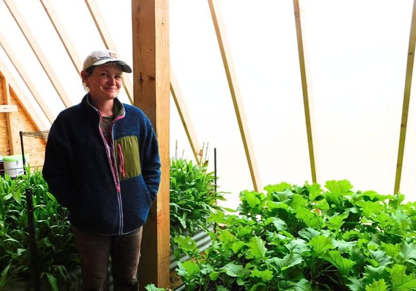A woman in a ball cap and light jacket stands inside a greenhouse surrounded by green, leafy plants.