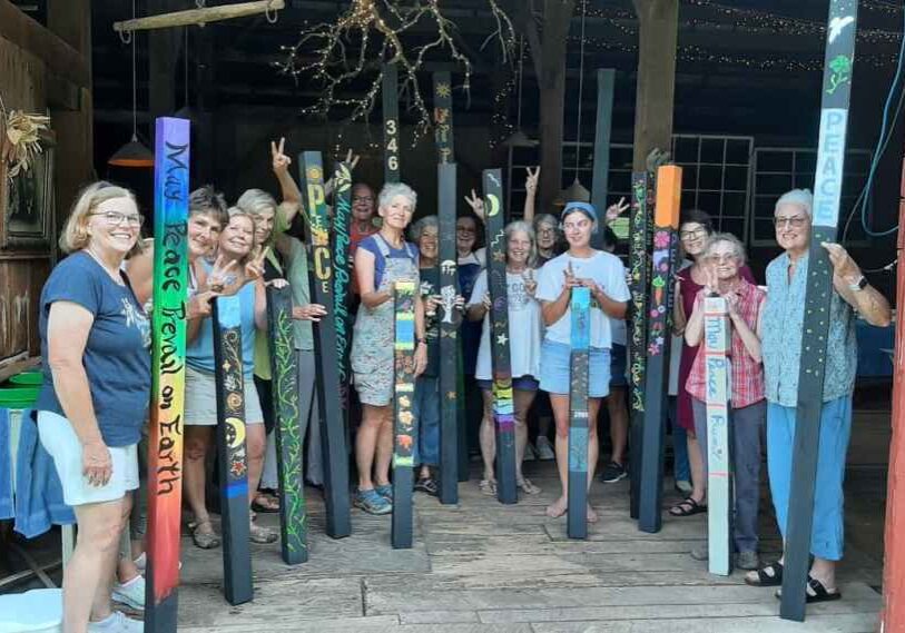 A group of people, mostly women of multiple ages stand in a rustic barn. Each one is holding a tall painted pole.