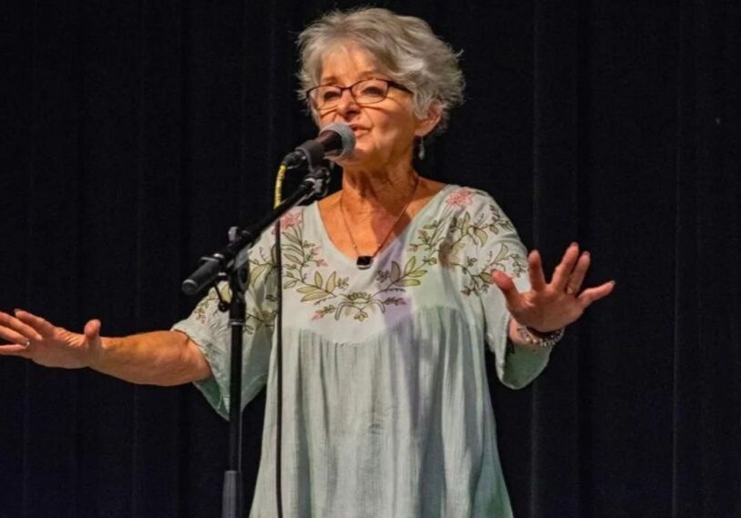 PHOTO-1-Power-of-Storytelling-by-Sandy-Webb-May-2025 An older woman with short gray hair stands behind a microphone on a stand and in front of a black curtain. Her arms are outstretched and her hands are flat with fingers spread widely.